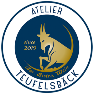 Das Logo des Atelier Teufelsbäck ist kreisrund. Der Hintergrund ist ein dunkles Blau. Die Schriftzeichen des Namens und die Beschreibung "Bar-Bistro-Weinstube" sind golden und in der Mitte kann man eine Ziege sehen.