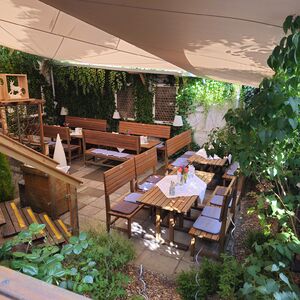 Auf dem quadratischen Foto ist der Biergarten des Atelier Teufelsbäck zu sehen. Die weißen Wände sind grün bewachsen mit Efeu und Wein. Die Bänke sind aus hellbraunem Naturholz, ebenso wie die Tische. Der Biergarten ist mit einem Sonnensegel überdacht. Das Geländer ist ebenfalls aus Holz und mit Glasplatten verstärkt.